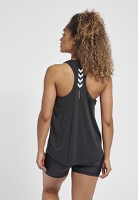 Frau mit lockigem Haar in schwarzem ärmellosem Sporttop mit weißem Chevron-Design und passenden schwarzen Shorts, rückwärts gewandt.