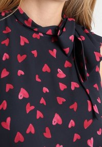 Marineblouse met kraag en strikdetail, voorzien van een patroon van kleine roze hartjes verspreid over de stof. Gladde textuur.