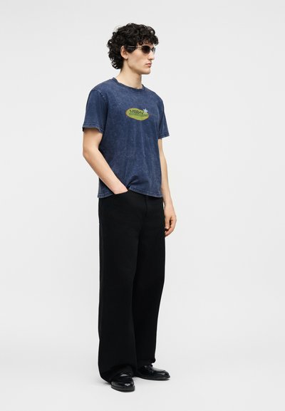 Νέος με σγουρά μαλλιά, που φοράει σκούρα γυαλιά ηλίου, μπλε φθαρμένο γραφικό μπλουζάκι, φαρδιά μαύρα παντελόνια και μαύρα παπούτσια, στέκεται με το ένα χέρι στην τσέπη.