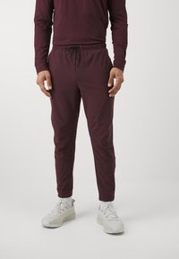 Burgundijas sporta bikses ar elastīgu jostu, auklu un sašaurinātām kājām, kopā ar vieglām baltām sporta apaviem un zeķēm.