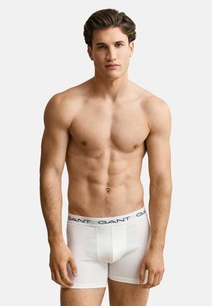 Ung mann med kort mørkt hår står, iført hvite GANT boxer briefs, mot en ensfarget lys bakgrunn.