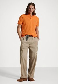 Πορτοκαλί πόλο πουκάμισο με γιακά, συνδυασμένο με ανοιχτό μπεζ παντελόνι με πιτσιλιές μπογιάς. Καφέ παπούτσια με υφή λεπτομέρειας.