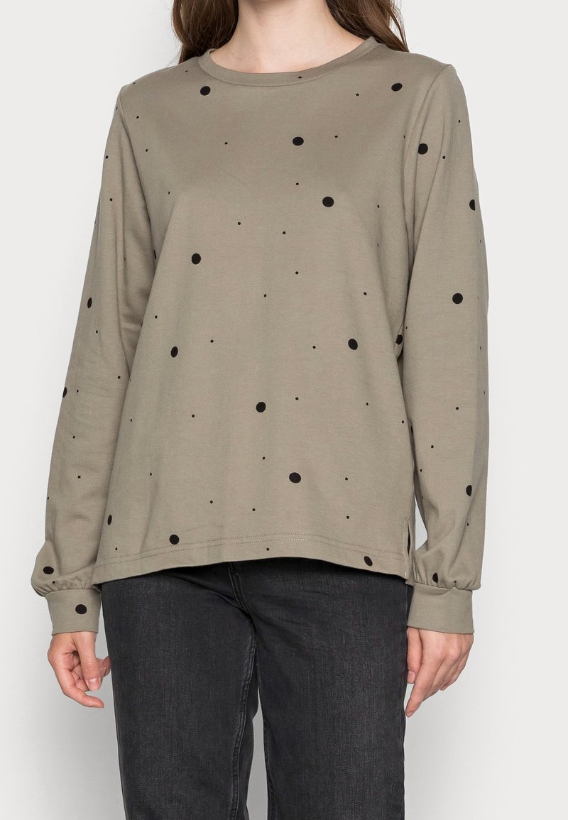 Chemise taupe à manches longues avec des petits et grands pois noirs dispersés, portée avec un jean en denim noir.