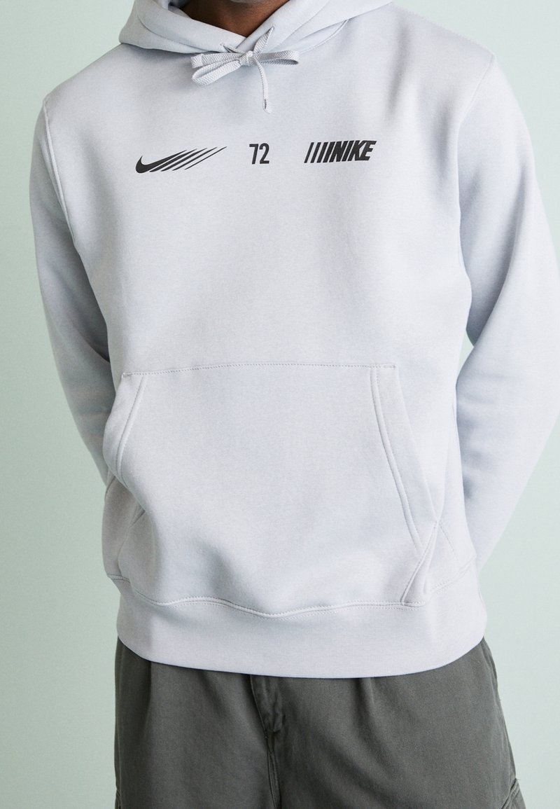 Světle šedá mikina s klokaní kapsou vpředu, kapucí na stažení a černými logy Nike umístěnými na hrudi. Měkká látka a uvolněný střih.