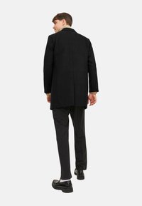 Schwarzer Wollmantel mit taillierter Passform, mittellang, mit einem Schlitz hinten, kombiniert mit schwarzen Hosen und gl�änzenden schwarzen Slippern.
