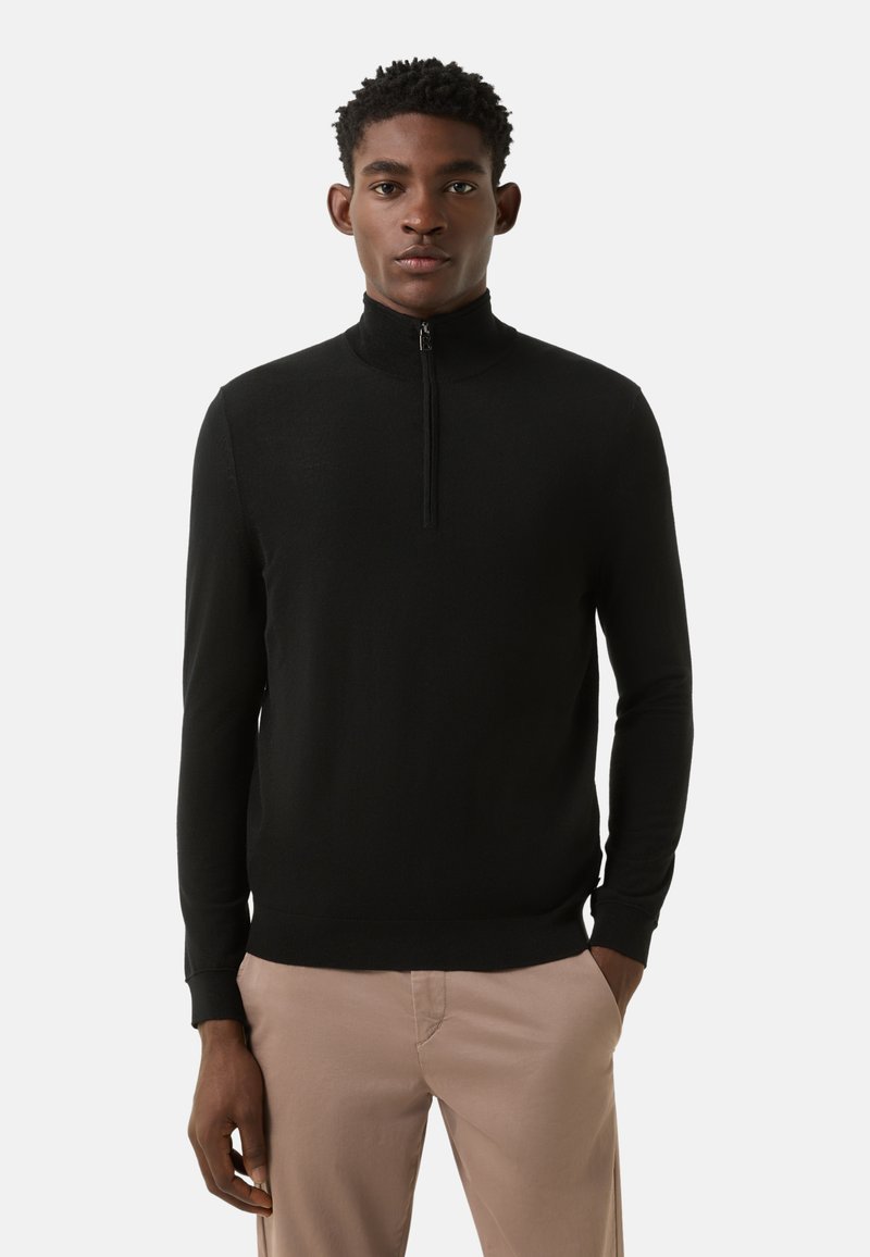 Zwart trui met rits en hoge kraag, gemaakt van fijn gebreid materiaal. Getailleerde mouwen en simpel, strak ontwerp. Model draagt lichte broek.