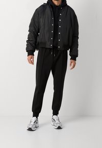 Svart kort bomberjacka med tryckknappar, bärs över en svart hoodie. Kombineras med svarta sweatpants och silver/vita löparskor.