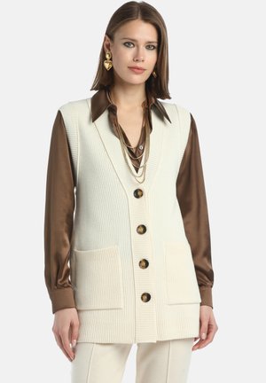 Crème ribgebreide top met v-hals en vier bruine knopen, twee voorzakken, gedragen over een bruine satijnen blouse met een gelaagde ketting.