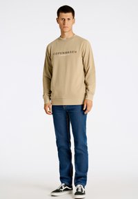 Beige sweatshirt med "COPENHAGEN" tryckt i mörkgrått, ihop med blå jeans och svarta sneakers. Långa ärmar och rundad halsringning.