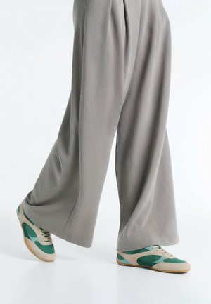 Personne portant un pantalon gris ample et des baskets vertes, beiges et bleues marchant sur un fond blanc.