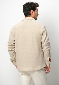Camicia beige strutturata con colletto, maniche lunghe e spacchi laterali nei polsini. Il tessuto sembra intrecciato e leggero, adatto per un abbigliamento informale.