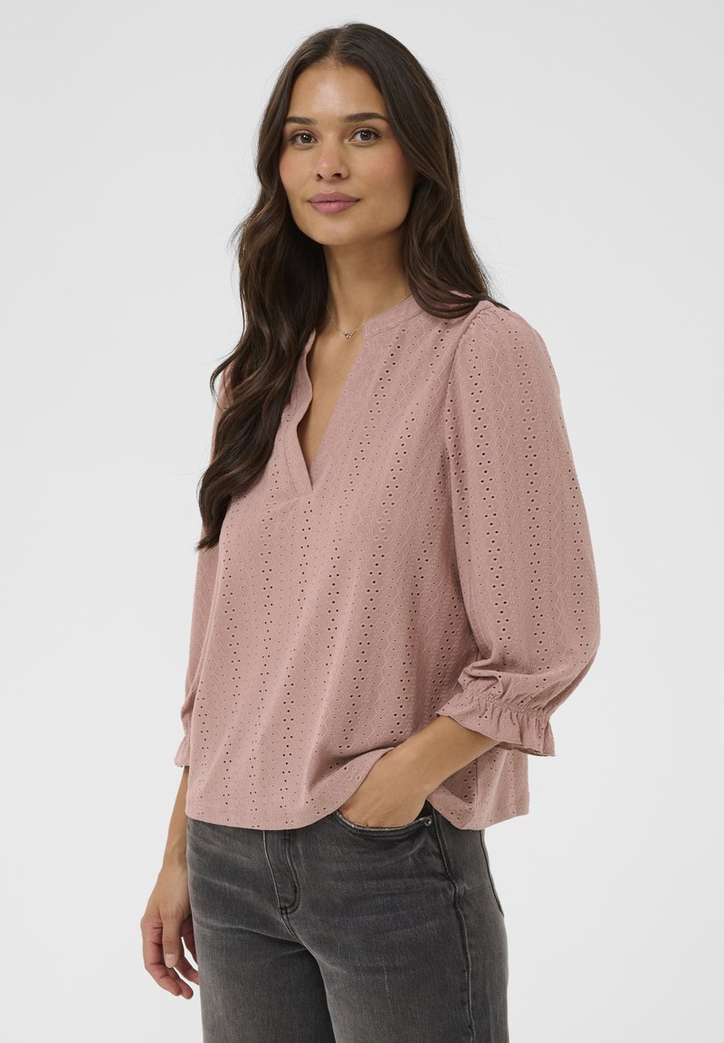 Blouse rose en œillets avec un col en V, manches 3/4 bouffantes, motif texturé et coupe décontractée, portée avec un jean foncé.