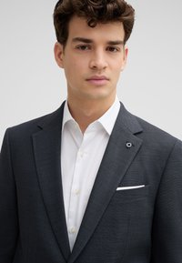 Blazer grigio scuro con design strutturato, rever a lancia e un emblema bianco e nero sul rever sinistro. Camicia bianca sotto.