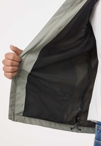 Hand die een open grijze jas vasthoudt en de zwarte mesh voering en verstelbare trekkoord aan de onderzoom toont.