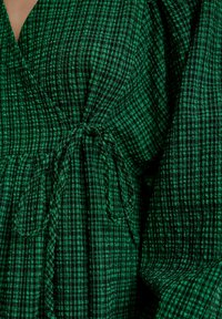 Groen geruite wikkeljurk met een gestructureerde stof, voorzien van een striksluiting en vloeiende mouwen. Verticale en horizontale patronen aanwezig.