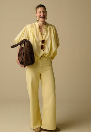 Femme en blouse jaune ample et pantalon à jambes larges riant, tenant un sac imprimé léopard et portant des lunettes de soleil sur la blouse.