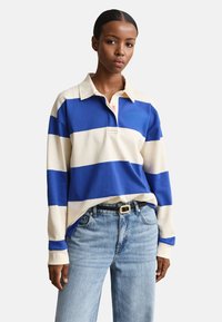 Femme portant un maillot de rugby à manches longues avec de larges rayures horizontales bleues et blanches, rentré dans un jean bleu clair avec une ceinture noire.