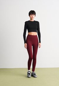 Kvinna som står mot en vit vägg iförd en svart långärmad croptop, vinröda högmidjade leggings, vita strumpor och silverskor.