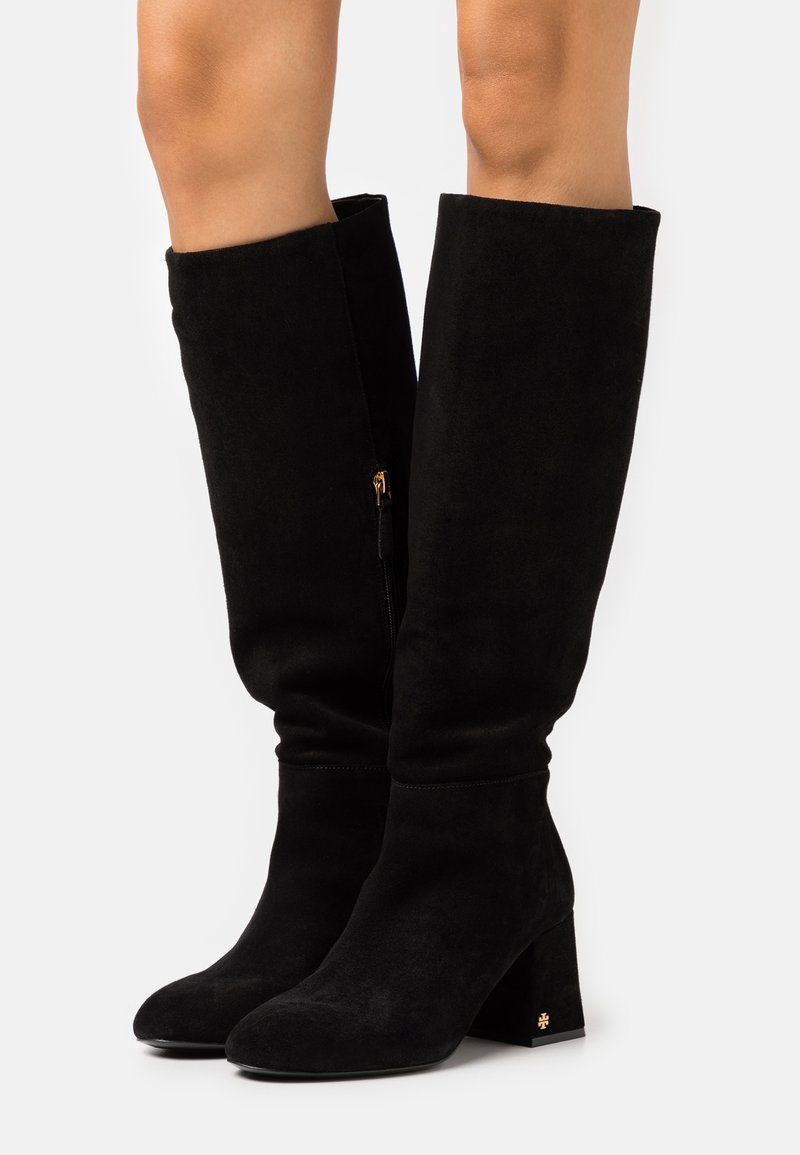 Bottes hautes jusqu'au genou en suède noir avec bout arrondi, talon épais, et fermeture éclair latérale avec garniture dorée. Texture lisse et design ajusté.