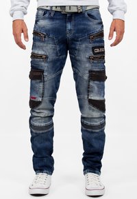 Pantalons cargo en denim bleu délavé avec des accents en tissu noir. Comprend plusieurs poches zippées et une coupe ajustée. Accents sur les cuisses.
