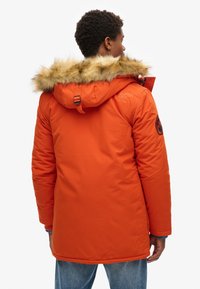 Chaqueta acolchada naranja con capucha forrada de piel sintética, que incluye puños acanalados, ajuste con cordón y logotipo en la parte posterior.