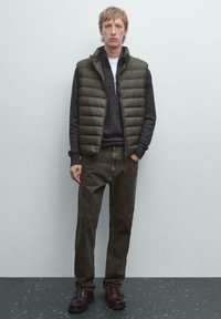 Ung mand iført en olivengrøn polstret vest, mørkegrå sweater med lynlås op til kvart, falmede sorte jeans og mørkebrune lædersko, står oprejst.