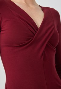 Top bordeaux a maniche lunghe con scollatura a V profonda e dettaglio a torsione sul seno, realizzato in tessuto elasticizzato con una superficie liscia.