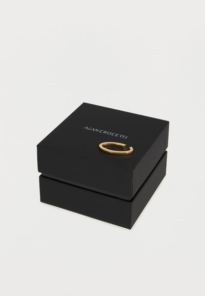 Μαύρο τετράγωνο κουτί κοσμημάτων με ασημένιο κείμενο λογότυπου και ένα απλό χρυσό ανοιχτό κρίκο σκουλαρικιού στην κορυφή, τοποθετημένο σε λευκή επιφάνεια.