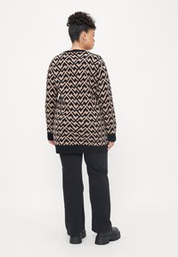 Knit cardigan with a geometric brown and black pattern, long sleeves, and black trim. Worn over black pants and chunky-soled shoes.