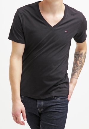 Mann trägt ein schwarzes V-Ausschnitt-T-Shirt mit kleinem Logo und dunkle Jeans, mit einem detaillierten Eulentattoo am linken Unterarm.