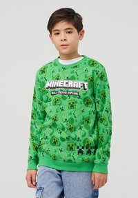 Felpa verde a tema Minecraft con un motivo ripetuto di facce di Creeper e elementi del gioco, collo a giro e polsini a costine.
