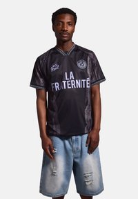 Jersey negro con "LA FRATERNITE" en azul claro. Presenta un cuello en V, patrones contrastantes y mangas cortas. Combinado con pantalones cortos ligeros deslavados en azul claro.