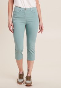Pantaloni capri azzurri chiari con due tasche frontali e un design aderente, abbinati a sneakers marroni e teal. Tessuto liscio con una leggera elasticità.