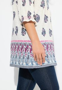 Hvid tunika med marineblå blomsterprint og lyserøde accenter, med et mønstret kant og flæseærmer. Kombineret med mørke denim jeans.