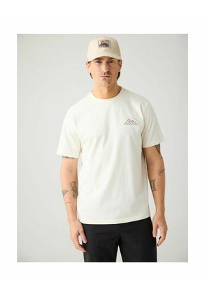 Man die een beige pet en een wit T-shirt draagt met een klein ontwerp van een berg en zon, staand tegen een effen lichte achtergrond.