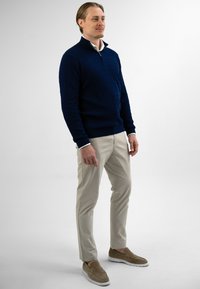 Man in een marineblauwe kabeltrui, witte overhemd, beige broek en bruine instappers, staande tegen een witte achtergrond.
