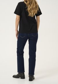 Černé tričko s krátkým rukávem, tmavě modré džíny se rovnými nohavicemi, kožené kotníkové boty s přezkami, modelka otočená zády na světlém pozadí.