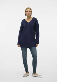 Marineblaues Oversized-Sweatshirt mit V-Ausschnitt, geripptem Kragen und langen Ärmeln, kombiniert mit hellblauen Jeans und silbernen Ballerinas.