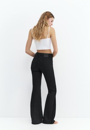 Donna con lunghi capelli mossi che indossa un top corto bianco e jeans neri a vita alta a zampa d'elefante, in piedi a piedi nudi su sfondo bianco.