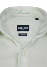 Wit katoenen overhemd met een moderne pasvorm. Voorzien van een ronde kraag, knoopsluiting en "Amour" geborduurd in lichtgroen aan de binnenkant van de kraag.