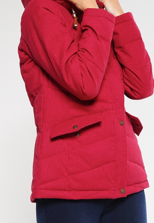 Personne portant une veste matelassée rouge avec des poches à rabat boutonnées et un pantalon bleu marine, ajustant le col de la veste avec les mains.