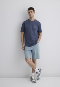 Blue cotton t-shirt with a round neck and logo detail, paired with light denim shorts. Worn with gray and silver sneakers and white socks.