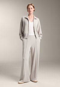 Ljusgrå zip-up jacka med sänkt axel, tillsammans med vida byxor i matchande färg. Vit åtsittande linne under, mjuk tygtextur.