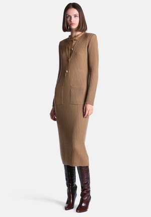 Femme portant une longue robe côtelée couleur camel avec des poches avant et des boutons dorés, associée à des bottes à talons hauts marron foncé.