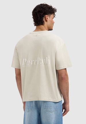 Man met krullend haar die een beige oversized T-shirt draagt met de tekst "PurePath" op de rug en lichtblauwe spijkerbroek, met de rug naar de kijker toe.