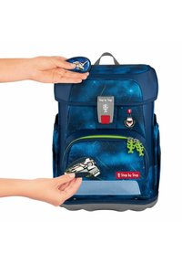 Zaino blu navy con motivo stellato. Presenta una tasca anteriore con cerniera verde, toppe personalizzabili e fondo grigio. Mano che posiziona una toppa.