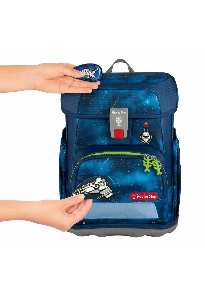 Zaino blu navy con motivo stellato. Presenta una tasca anteriore con cerniera verde, toppe personalizzabili e fondo grigio. Mano che posiziona una toppa.