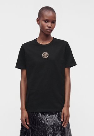 Femme aux cheveux courts portant un T-shirt noir avec un petit emblème circulaire doré au centre et une jupe noire brillante, debout sur un fond blanc.