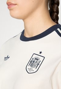 Biała koszulka sportowa z granatowym kołnierzykiem i trzema paskami. Zawiera granatowy emblemat hiszpańskiej federacji piłkarskiej oraz gwiazdkę nad nim.