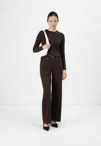 Braunes Langarmshirt kombiniert mit braunen weit geschnittenen Jeans im Leopardenmuster. Modell hält eine weiße Tasche und trägt schwarze Lackschuhe.